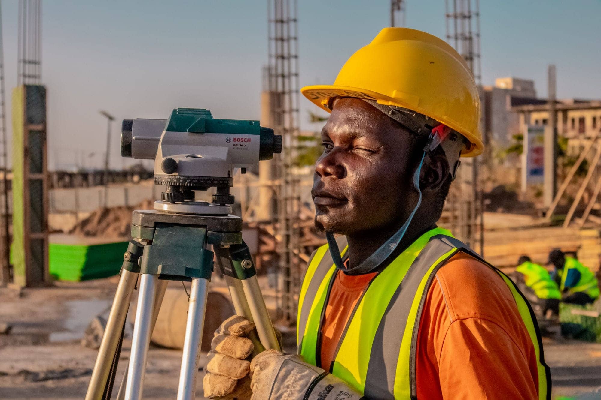 Site worker in a yellow hard hat and safety vest beside a surveying level on a tripod at a construction site