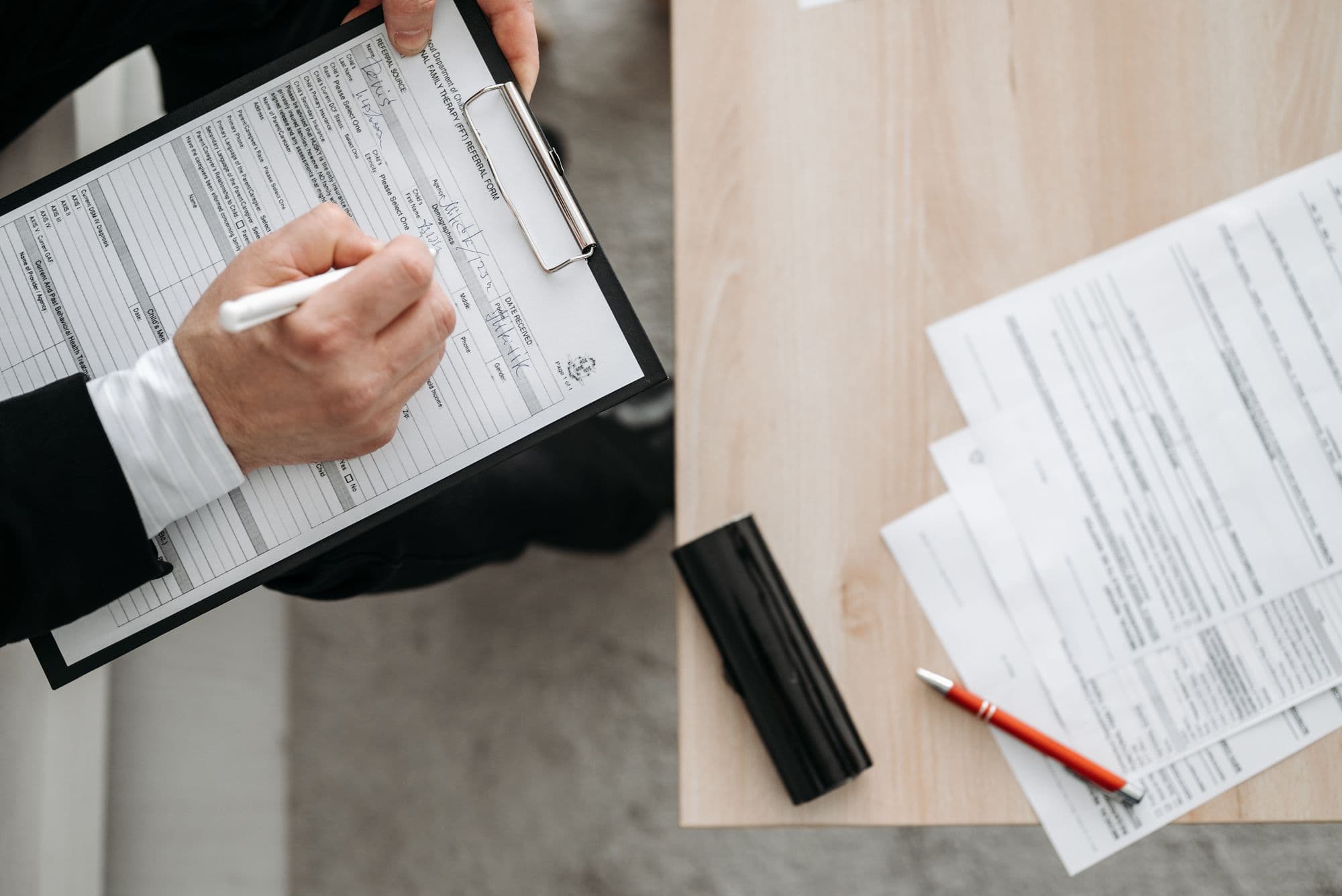 Overhead view of a person filling in a form on a clipboard beside printed documents and a pen