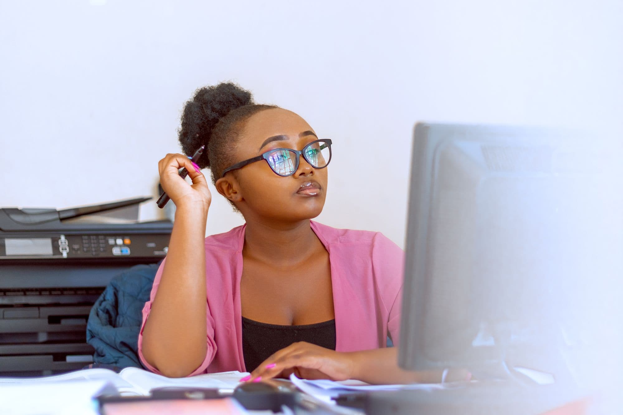 Young woman in glasses working at a desktop computer, with a printer and paperwork on the desk beside her