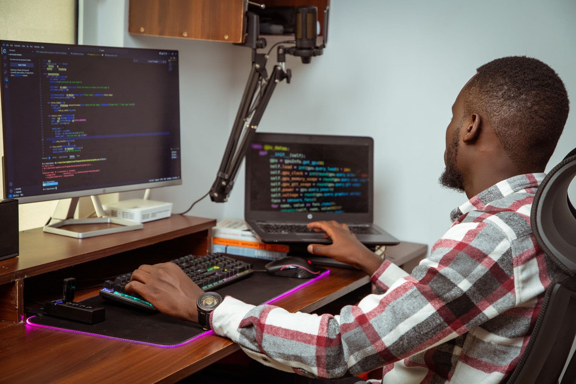 Man in a checked shirt working at a desk with a desktop monitor and a laptop