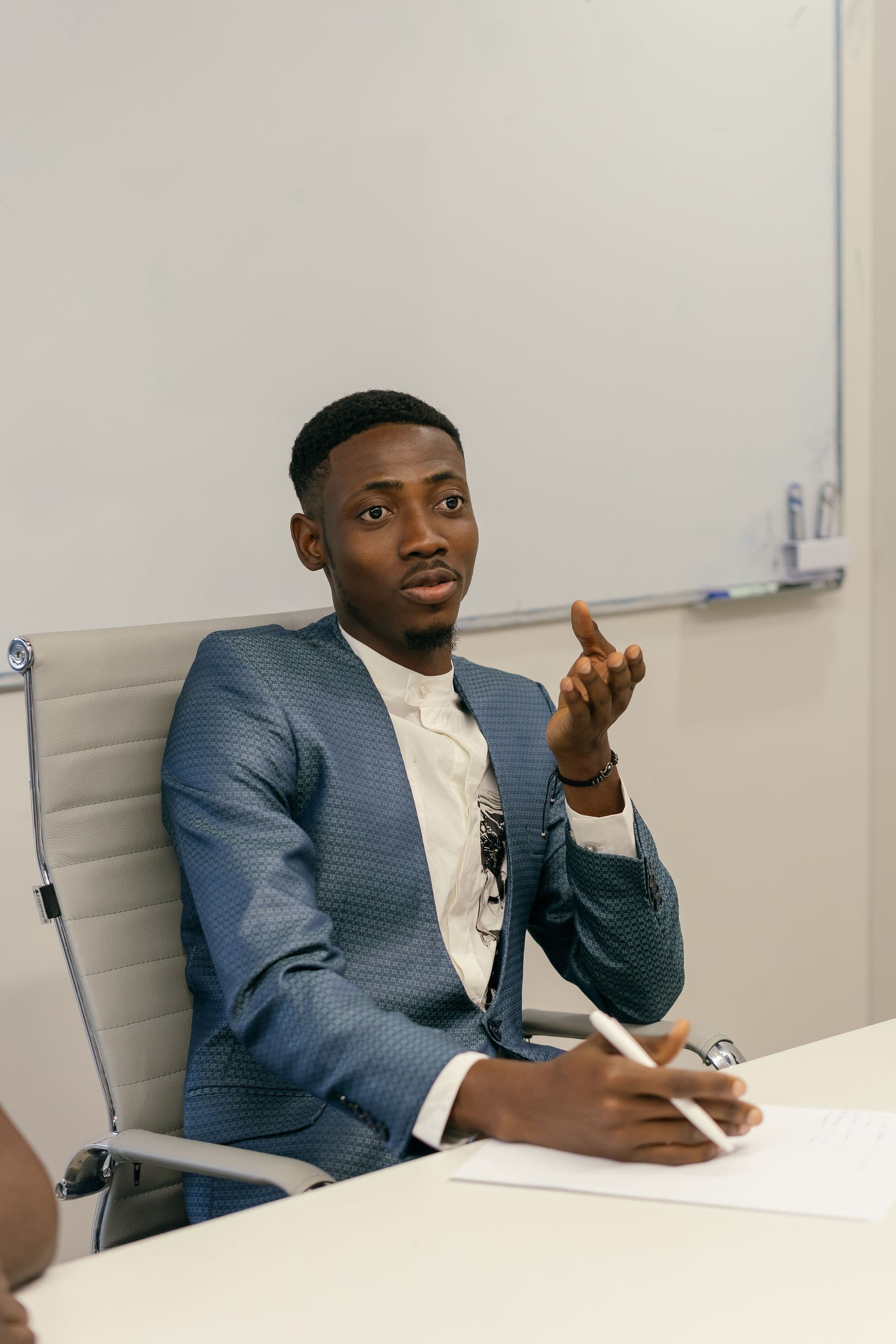 Young man in a blue blazer speaking across a table during a meeting, pen and paper in hand