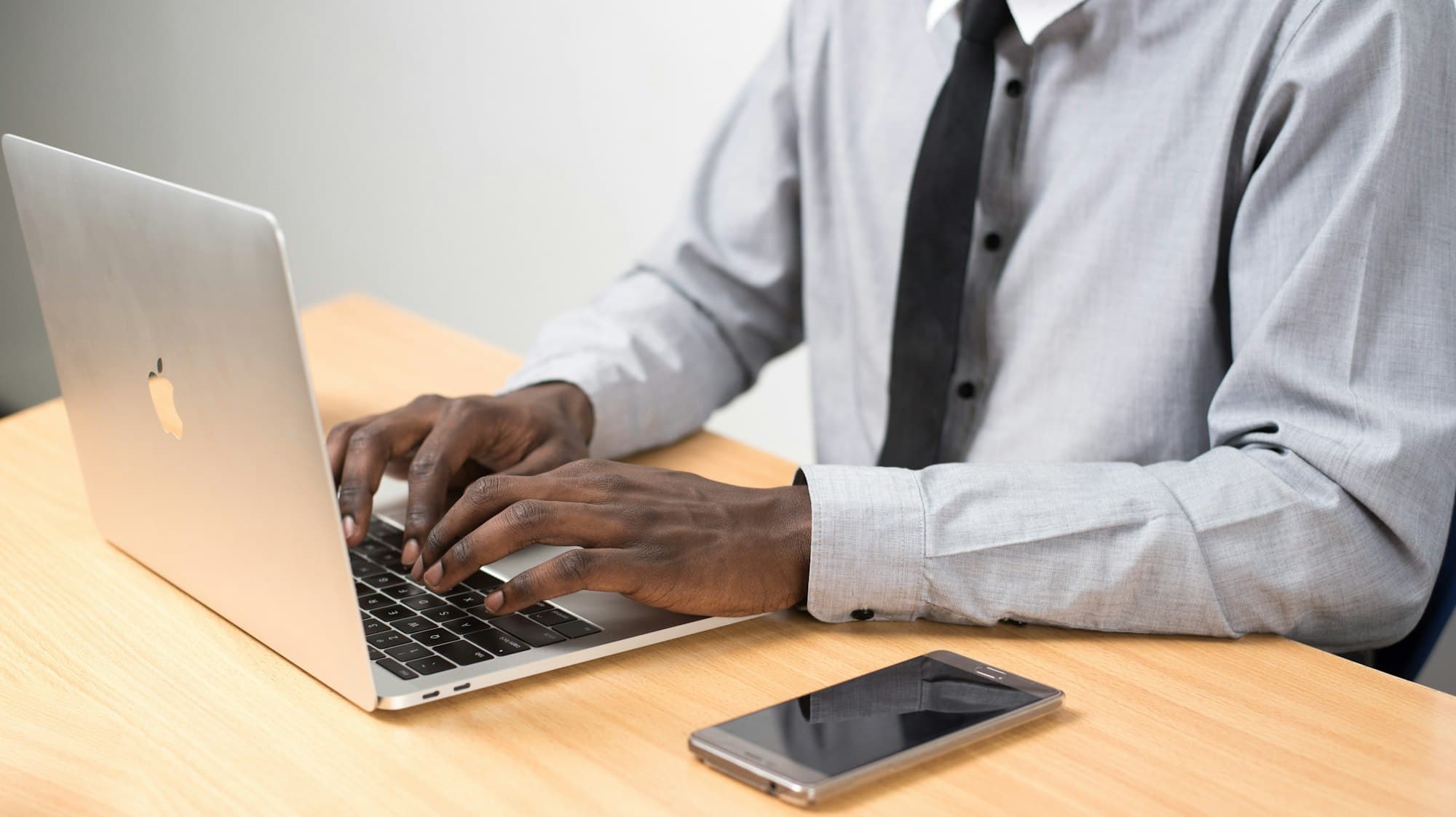 Close-up of a man in a shirt and tie typing on a laptop, with a smartphone on the desk